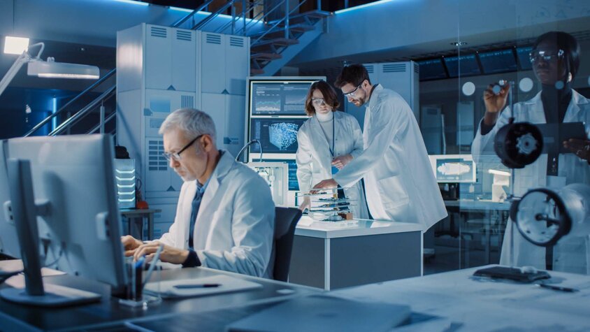 A diverse team of industrial scientists and engineers in white lab coats is working diligently in a research laboratory. They are using computers and communicating with each other while developing heavy machinery. In the background, technical drawings are visible, highlighting the complexity of their projects. The scene radiates professionalism and teamwork, with a focus on innovation in forming technology and mechanical engineering.