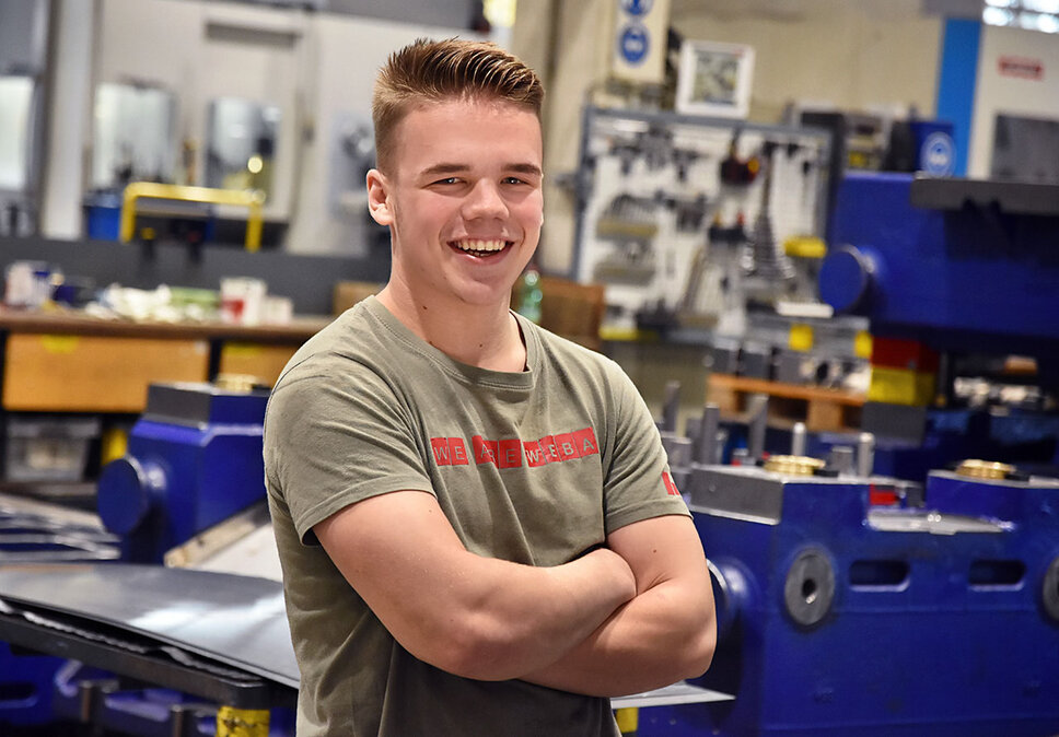 Ein Lehrling bei weba Werkzeugbau, fotografiert in der Produktionshalle, lächelt direkt in die Kamera. Er trägt ein T-Shirt mit der Aufschrift 'We are WEBA', was seinen Stolz und seine Zugehörigkeit zum Unternehmen zum Ausdruck bringt. Im Hintergrund sind mehrere Werkbänke und Teile eines dunkelblauen Umformwerkzeugs sichtbar, was auf die praktische und lernintensive Umgebung hinweist, in der der Lehrling ausgebildet wird. Das Bild strahlt Jugendlichkeit, Optimismus und das Engagement des Lehrlings für seine Ausbildung aus, während es gleichzeitig die unterstützende und lehrreiche Atmosphäre bei weba hervorhebt.