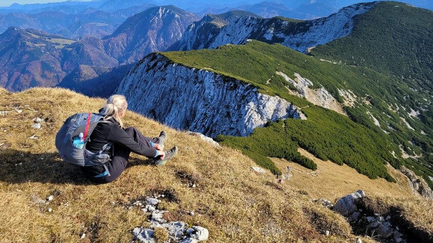 Freiheit, Reflexion und neue Perspektiven Bettina Gmainer mit Rucksack sitzt auf einer grasbewachsenen Anhöhe und blickt auf eine beeindruckende Berglandschaft mit grünen und felsigen Gipfeln.
