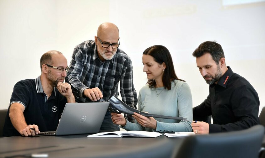 Drei männliche und eine weibliche Mitarbeiterin von weba Werkzeugbau sitzen in einer Besprechung über ein Stahlbauteil. Ein Laptop und Unterlagen liegen auf dem Tisch.