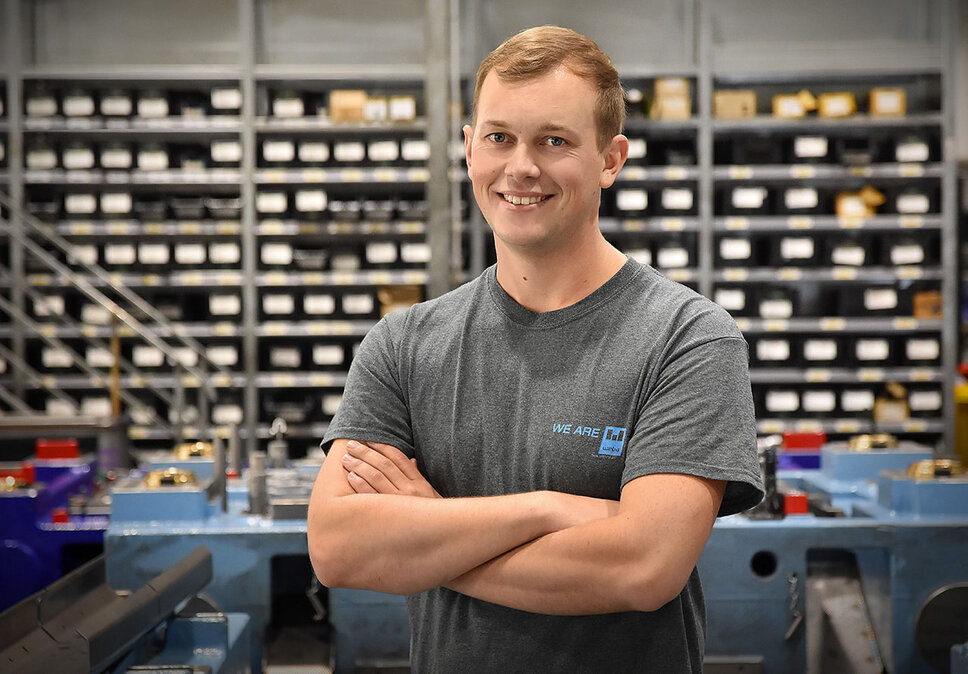 Ein Werkzeugbautechniker von weba Werkzeugbau, fotografiert in der Produktionsumgebung, blickt direkt und mit einem stolzen Lächeln in die Kamera. Er steht in einer selbstbewussten Pose, die seinen Stolz auf seine Arbeit unterstreicht. Im Hintergrund sind ordentlich angeordnete Betriebsmittelregale und ein markantes blaues Umformwerkzeug zu sehen, was auf die gut organisierte und technologisch fortschrittliche Arbeitsumgebung hinweist. Das Bild vermittelt ein Gefühl von Professionalität und Zufriedenheit im Arbeitsumfeld, und hebt die persönliche Verbindung des Technikers zu seiner Arbeit und seinem Arbeitsplatz hervor.