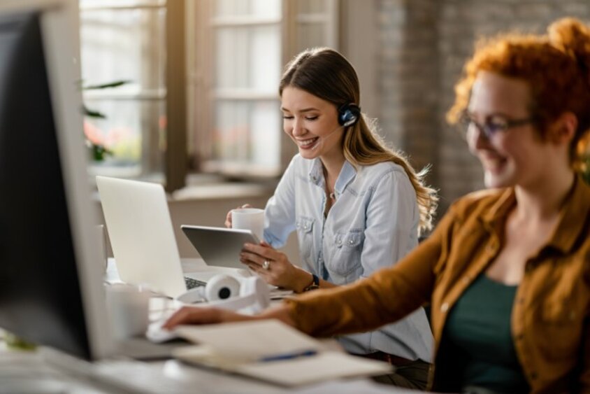 Lächelnde weiblich gelesene Person mit Headset, die Kaffee trinkt und im Büro das Touchpad nutzt.