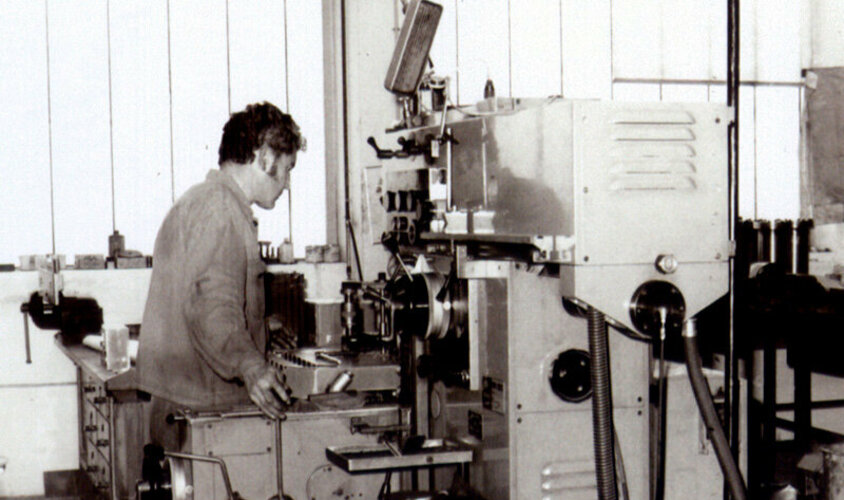  A black-and-white photo from the 1980s shows a machining technician operating a conventional drilling machine.