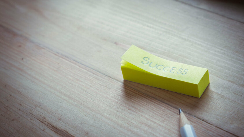 Best Practice bei weba – Erfolg durch gemeinsame Ideen Gelber Notizzettel mit dem Wort „Success“ auf Holztisch, daneben ein Bleistift.