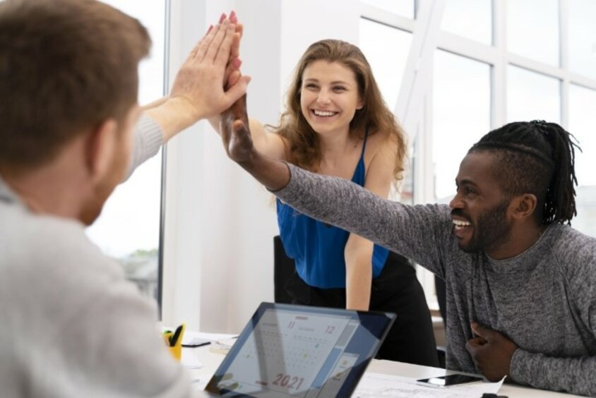 Drei Personen sitzen lächelnd an einem Besprechungstisch und geben sich High-Five.