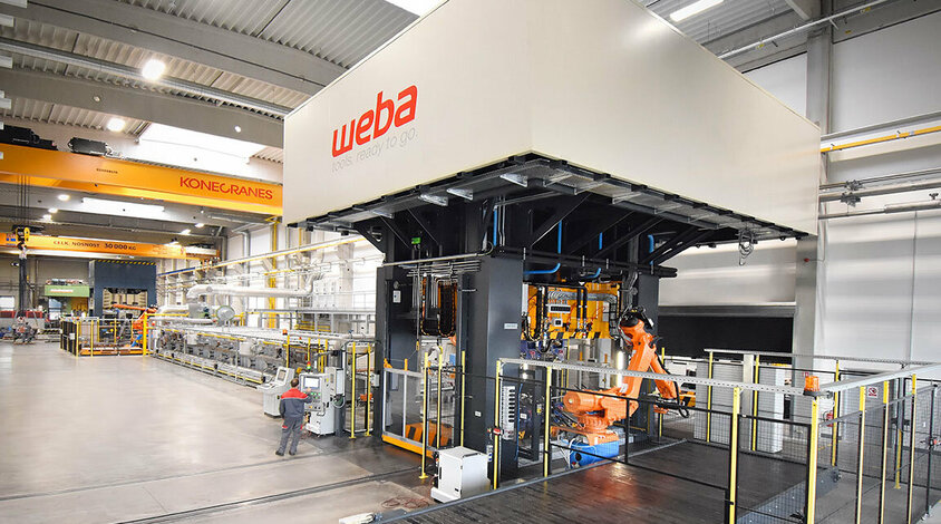 A view of a series production line for hot forming, focusing on the forming press and the conveyor belt transporting finished components. A press operator stands next to the control panel, intently monitoring the process. In the background, a portion of the roller hearth furnace is visible, used for heating the metal parts prior to forming. The forming press is utilized for both series production and tool tryouts.