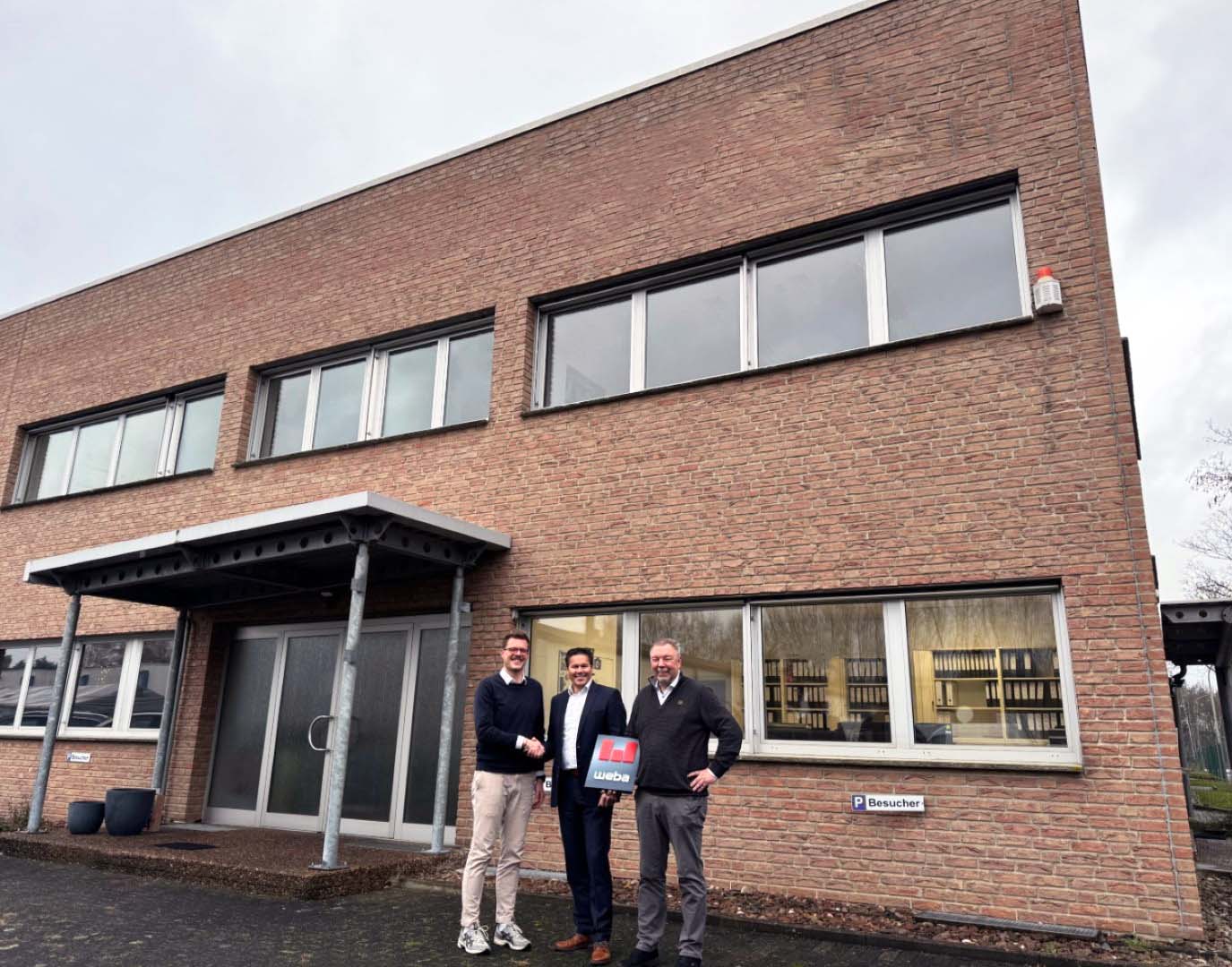 Three men standing in front of a brick building, two shaking hands while the third holds a document with weba branding.